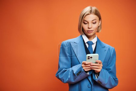 Foto de Mujer joven positiva con el pelo corto rubia posando con su teléfono inteligente sobre fondo naranja - Imagen libre de derechos