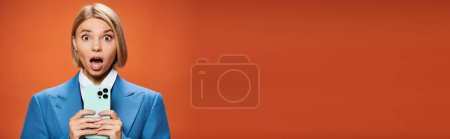 Foto de Sorprendió a la joven con el pelo corto rubia posando con su teléfono inteligente sobre fondo naranja, pancarta - Imagen libre de derechos