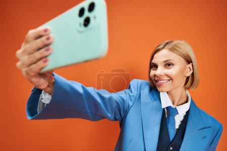 Foto de Feliz mujer bien vestida con pelo corto y rubio en traje elegante sosteniendo el teléfono sobre fondo naranja - Imagen libre de derechos