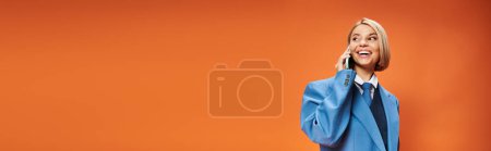 Foto de Alegre mujer elegante con pelo corto y rubio en traje elegante sosteniendo el teléfono sobre fondo naranja, pancarta - Imagen libre de derechos