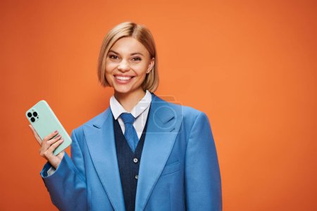 Foto de Mujer sofisticada positiva con pelo corto y rubio en traje elegante sosteniendo el teléfono sobre fondo naranja - Imagen libre de derechos