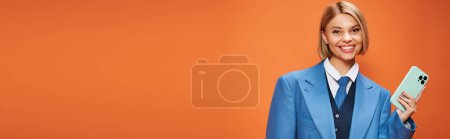 Foto de Mujer elegante feliz con pelo corto y rubio en traje elegante sosteniendo el teléfono sobre fondo naranja, pancarta - Imagen libre de derechos