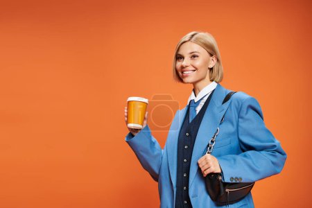 Foto de Mujer sofisticada alegre con pelo corto rubio sosteniendo bolso y café sobre fondo naranja - Imagen libre de derechos