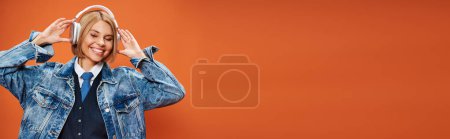 Photo for Jolly woman with blonde hair with headphones in denim jacket posing on orange backdrop, banner - Royalty Free Image