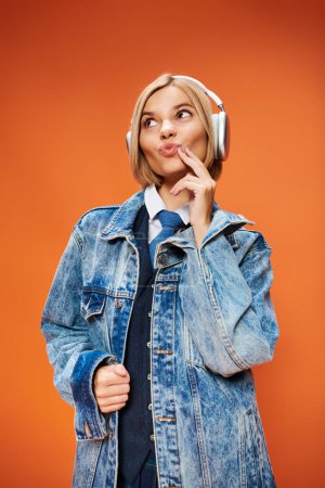 Photo for Joyous young woman with blonde hair with headphones in denim jacket posing on orange backdrop - Royalty Free Image