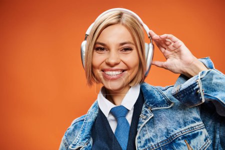 Photo for Joyful young woman with blonde hair with headphones in denim jacket posing on orange backdrop - Royalty Free Image