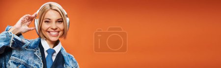 Foto de Alegre joven con el pelo rubio con auriculares en chaqueta de mezclilla posando sobre fondo naranja, pancarta - Imagen libre de derechos