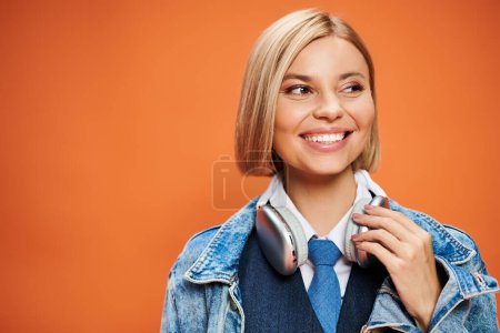 Téléchargez les photos : Joyeuse femme attrayante avec des cheveux blonds avec des écouteurs dans la veste en denim posant sur fond orange - en image libre de droit