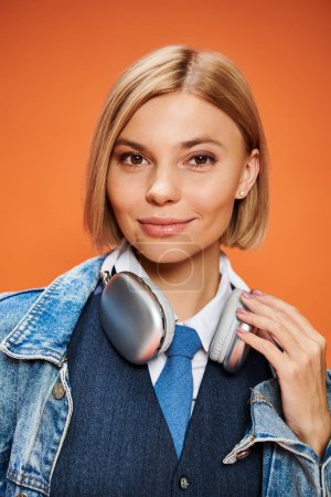 Photo for Positive stylish woman with blonde hair with headphones in denim jacket posing on orange backdrop - Royalty Free Image