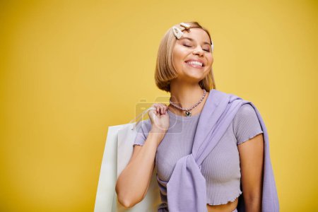 Foto de Mujer contenta con accesorios en traje elegante posando con bolsa de compras sobre fondo amarillo - Imagen libre de derechos