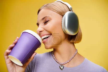 Foto de Mujer alegre y elegante con auriculares disfrutando de la música y el café caliente mientras posa en el fondo amarillo - Imagen libre de derechos