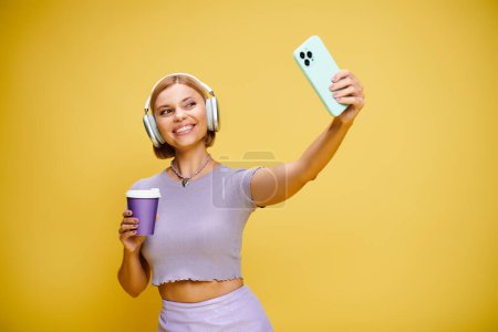 good looking cheerful woman with headphones and cell phone enjoying her coffee on yellow backdrop