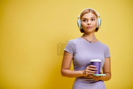 Foto de Joven mujer alegre con auriculares y teléfono celular disfrutando de su café sobre fondo amarillo - Imagen libre de derechos