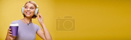 Foto de Mujer alegre con auriculares disfrutando de la música y el café caliente mientras posa sobre el fondo amarillo, pancarta - Imagen libre de derechos