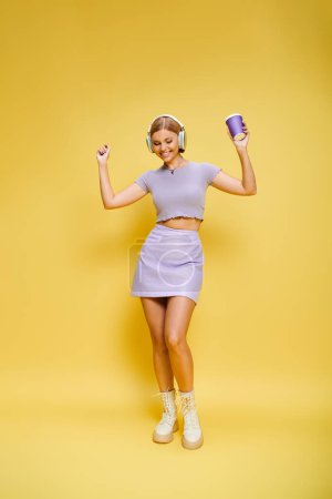 Foto de Mujer con estilo positivo con auriculares disfrutando de la música y el café caliente mientras posa en el fondo amarillo - Imagen libre de derechos