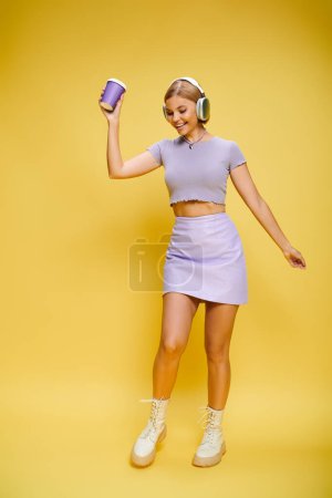 Foto de Mujer de moda alegre con auriculares disfrutando de la música y el café caliente mientras posa en el fondo amarillo - Imagen libre de derechos