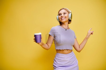 Foto de Alegre mujer de moda con auriculares disfrutando de la música y el café caliente mientras posa en el fondo amarillo - Imagen libre de derechos
