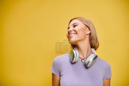 Foto de Exquisita mujer alegre con pelo corto y rubio y auriculares disfrutando de la música en el fondo amarillo - Imagen libre de derechos