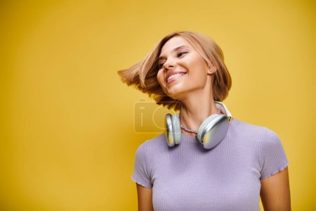 Téléchargez les photos : Femme joyeuse polie avec cheveux blonds courts et écouteurs profitant de la musique sur fond jaune - en image libre de droit
