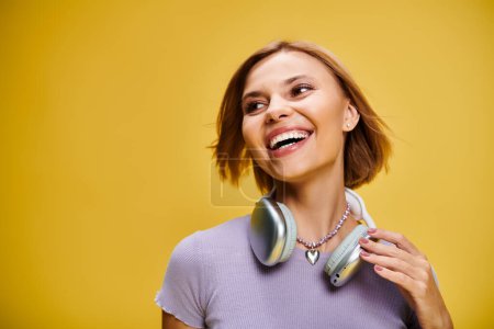 Téléchargez les photos : Gracieuse femme joyeuse avec cheveux blonds courts et écouteurs appréciant la musique sur fond jaune - en image libre de droit