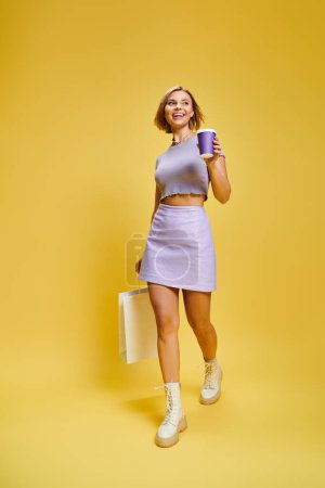 Photo for Joyful young woman in vibrant attire with short hair posing with coffee and shopping bag in hands - Royalty Free Image