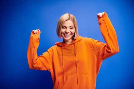 Foto de Mujer rubia alegre con el pelo corto en sudadera con capucha naranja vibrante posando activamente sobre fondo azul - Imagen libre de derechos