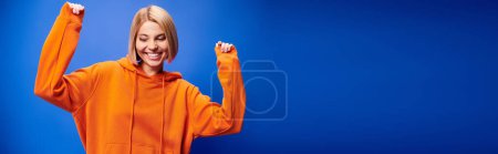 Foto de Mujer contenta con el pelo corto en sudadera con capucha naranja vibrante posando activamente sobre fondo azul, bandera - Imagen libre de derechos
