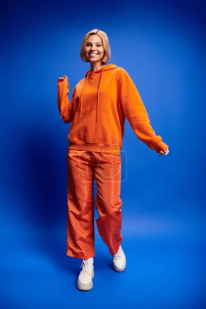 Foto de Mujer rubia contenta con el pelo corto en sudadera con capucha naranja vibrante posando activamente sobre fondo azul - Imagen libre de derechos