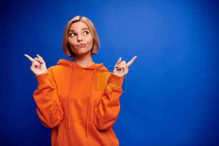 Photo for Glad blonde woman with short hair in vibrant orange hoodie posing actively on blue backdrop - Royalty Free Image