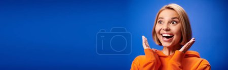 Foto de Mujer positiva con el pelo corto en sudadera con capucha naranja vibrante posando activamente sobre fondo azul, bandera - Imagen libre de derechos