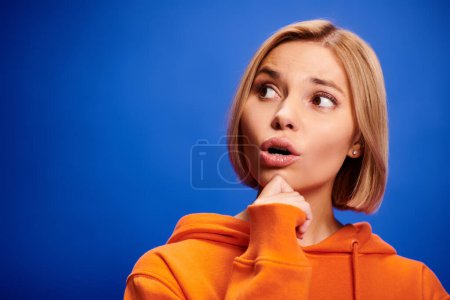 Foto de Atractiva mujer rubia desconcertada con pelo corto en sudadera con capucha naranja elegante posando sobre fondo azul - Imagen libre de derechos