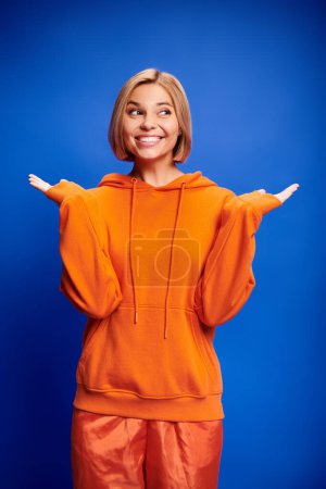 Foto de Hermosa mujer alegre con el pelo corto en sudadera con capucha naranja vibrante posando activamente sobre fondo azul - Imagen libre de derechos