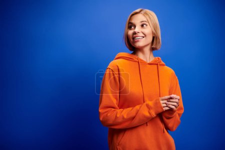 Foto de Atractiva mujer alegre con el pelo corto en sudadera con capucha naranja vibrante posando activamente sobre fondo azul - Imagen libre de derechos