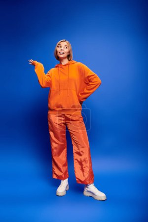 Foto de Atractiva mujer alegre con el pelo corto en sudadera con capucha naranja vibrante posando activamente sobre el telón de fondo azul - Imagen libre de derechos
