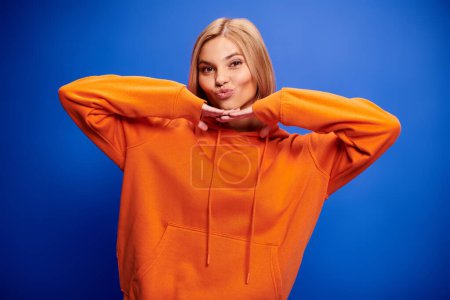 Foto de Mujer alegre elegante con pelo corto en sudadera con capucha naranja vibrante posando activamente sobre el telón de fondo azul - Imagen libre de derechos