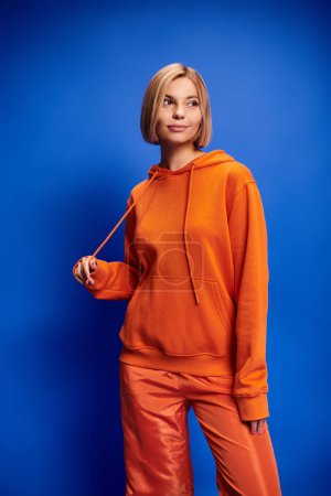 Photo for Exquisite cheerful woman with short hair in vibrant orange hoodie posing actively on blue backdrop - Royalty Free Image