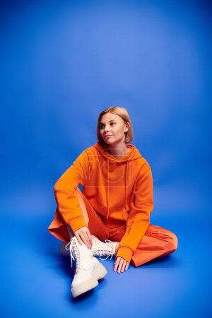 Photo for Debonair cheerful woman with short hair in vibrant orange hoodie posing actively on blue backdrop - Royalty Free Image