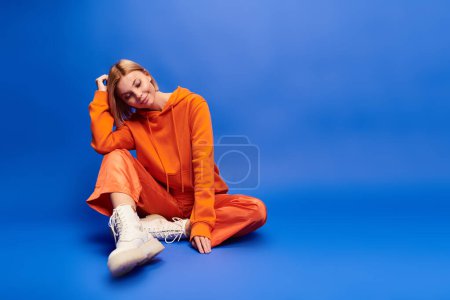 Foto de Mujer alegre de buen gusto con el pelo corto en sudadera con capucha naranja vibrante posando activamente sobre fondo azul - Imagen libre de derechos