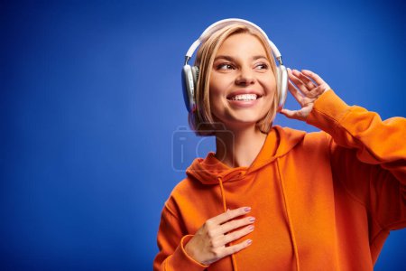Foto de Atractiva mujer alegre con pelo rubio corto y auriculares disfrutando de la música en el fondo azul - Imagen libre de derechos