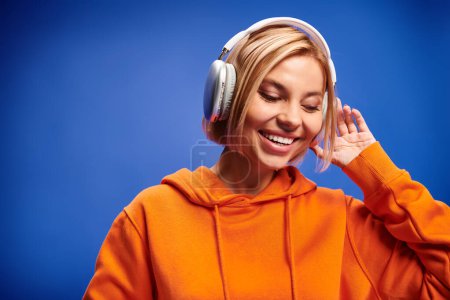 Foto de Mujer bastante elegante con pelo corto y rubio y auriculares disfrutando de la música en el fondo azul - Imagen libre de derechos