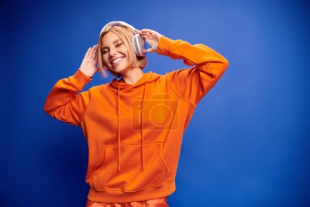 Foto de Mujer elegante de buen aspecto con pelo corto rubio y auriculares disfrutando de la música en el telón de fondo azul - Imagen libre de derechos
