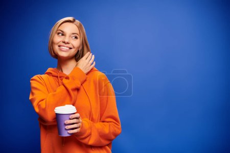 Foto de Atractiva mujer alegre con pelo corto y rubio en ropa vibrante sosteniendo taza de café sobre fondo azul - Imagen libre de derechos