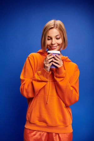 Foto de Hermosa mujer alegre con pelo corto y rubio en ropa vibrante sosteniendo taza de café sobre fondo azul - Imagen libre de derechos