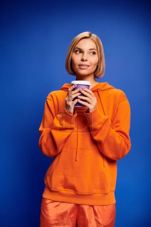 Foto de Atractiva mujer alegre con pelo corto y rubio en ropa vibrante sosteniendo taza de café sobre fondo azul - Imagen libre de derechos