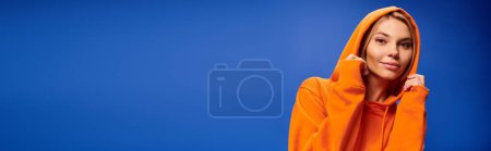 Photo for Pretty jolly woman with short hair in vivid attire with hood on posing on blue backdrop, banner - Royalty Free Image