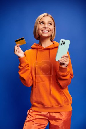 Foto de Hermosa mujer alegre con cabello rubio en sudadera naranja vibrante posando con tarjeta de crédito y teléfono - Imagen libre de derechos