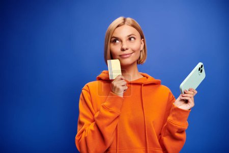 Téléchargez les photos : Séduisante femme joyeuse avec des cheveux blonds à capuche orange vif posant avec carte de crédit et téléphone - en image libre de droit