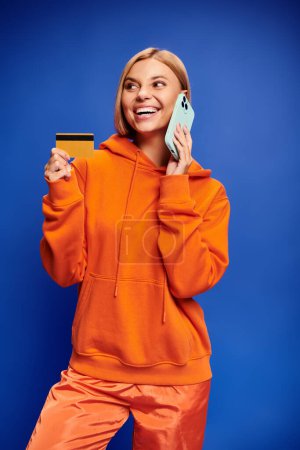 Foto de Seductora mujer alegre con el pelo rubio en sudadera naranja vibrante posando con tarjeta de crédito y teléfono - Imagen libre de derechos
