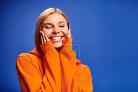 beautiful cheerful woman with hair in vibrant orange attire posing with closed eyes on blue backdrop