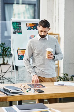 A man relaxes at a table with a cup of coffee in a bustling office environment, taking a moment to regroup amidst corporate hustle.